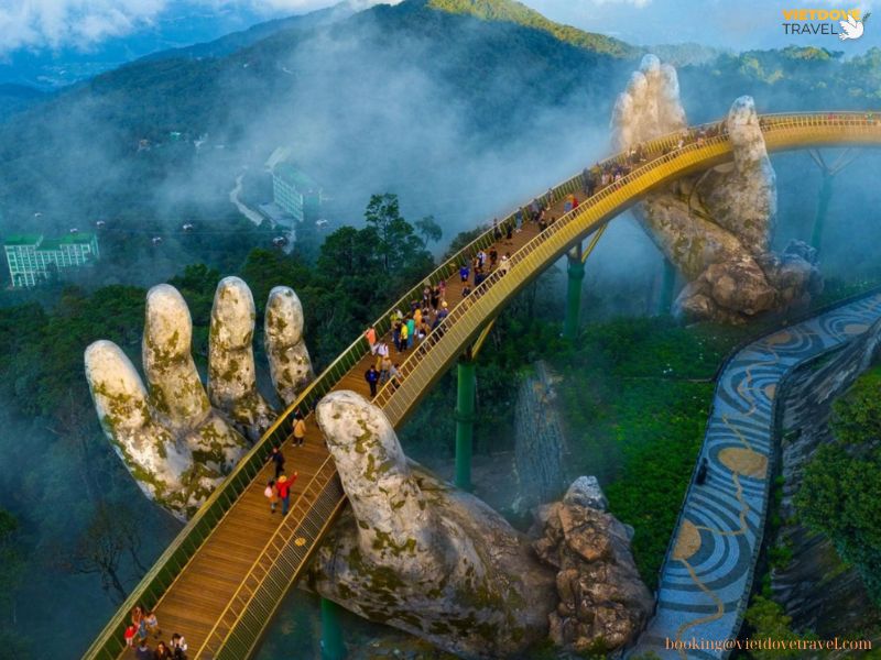 A dramatic photo of the Golden Bridge at Ba Na Hills, supported by giant stone hands with misty mountains in the background
